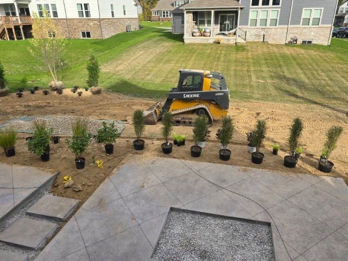 Skid steer landscaping in yard with new concrete patio. Trees in pots lined up, houses in background.