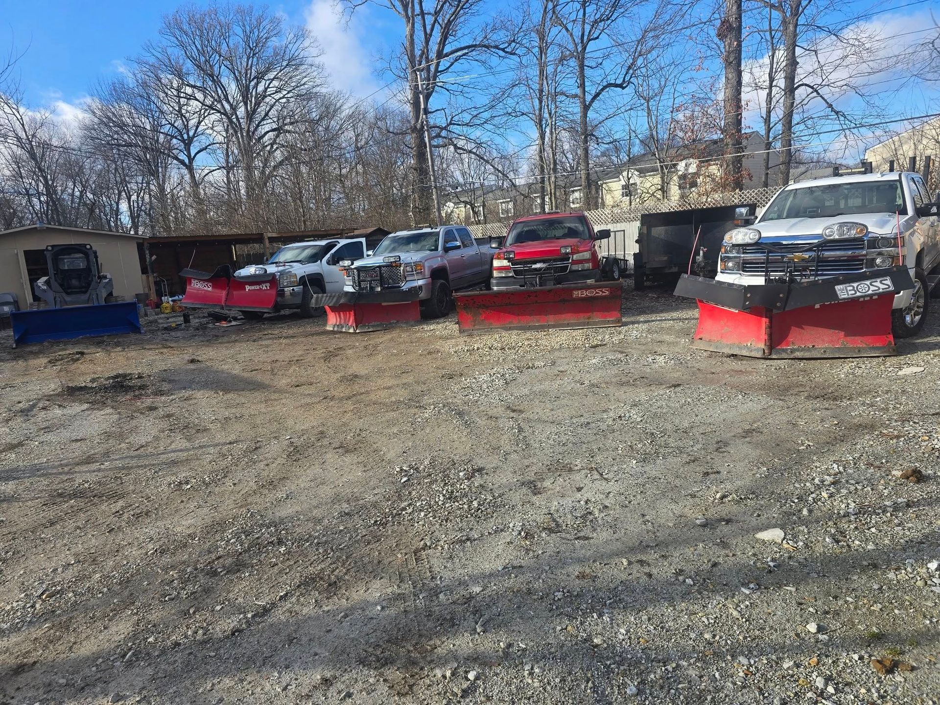 Trucks with snowplows parked on a gravel lot near trees.