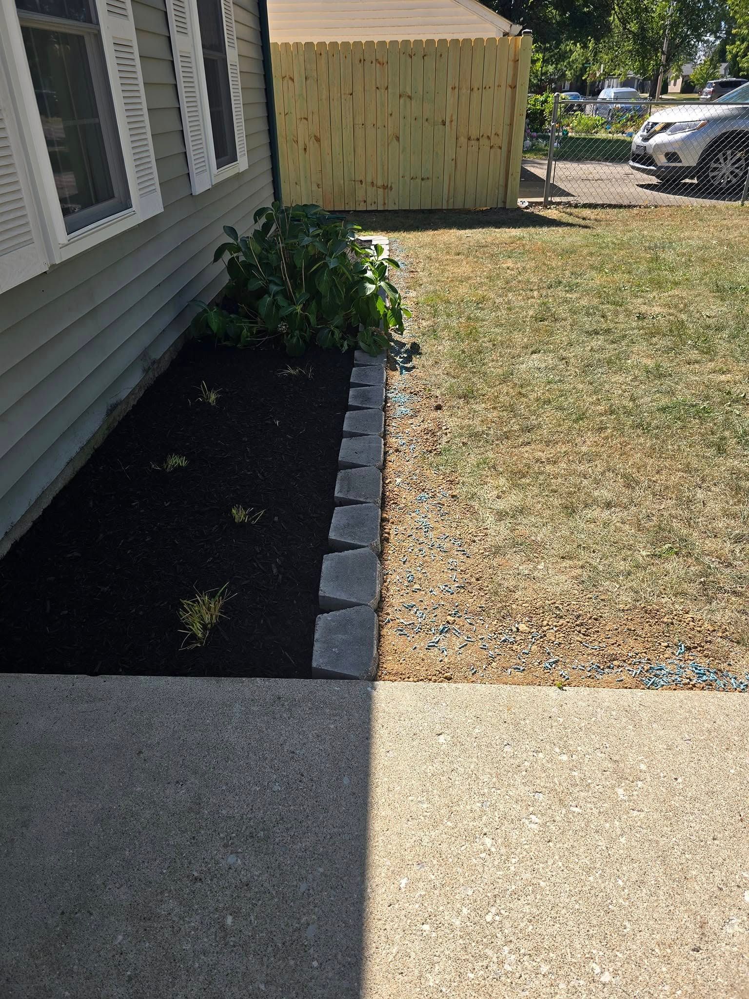 A house with a newly mulched flower bed bordered by gray bricks. The lawn is partly brown.