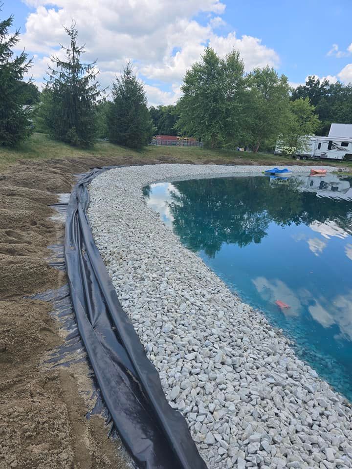 A pond with a gravel border and a black liner.  Trees and a cloudy sky are in the background.