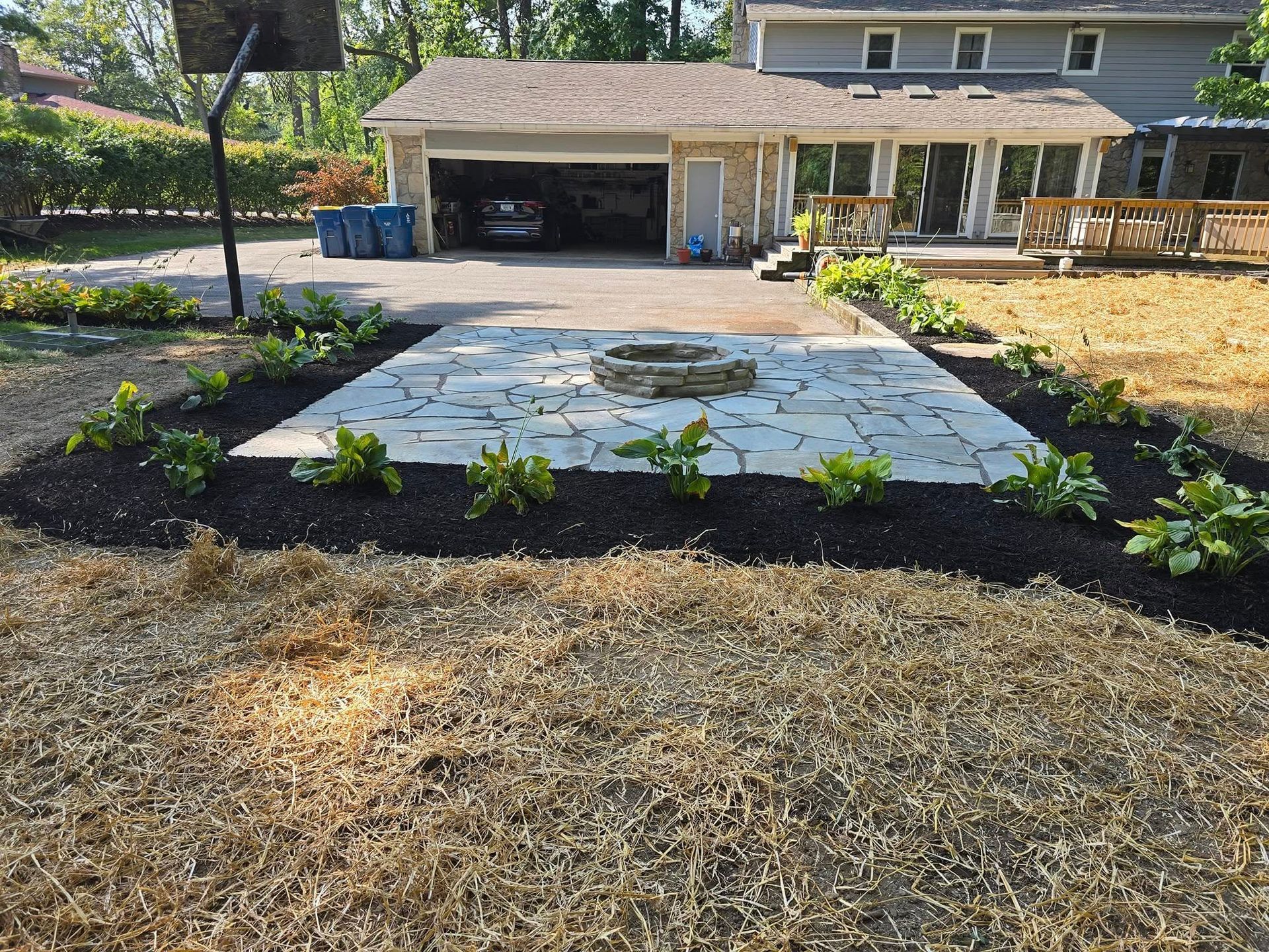 Backyard patio with fire pit, bordered by landscaping beds with green plants, house and driveway in background.