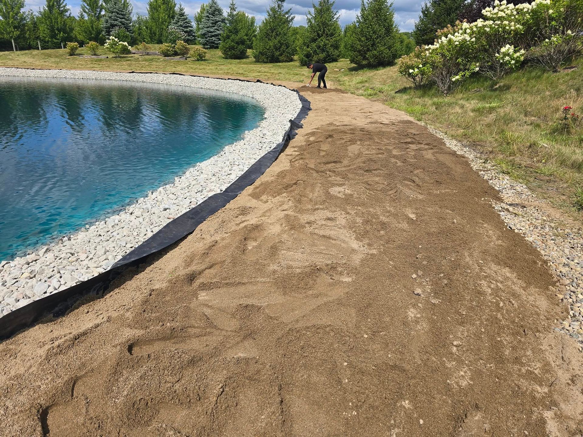 A pond with a stone border and a dirt path, a person stands in the background.