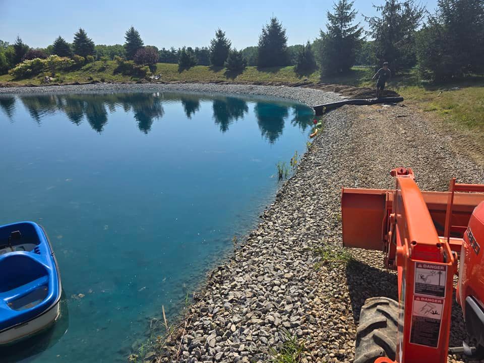 Blue pond with gravel shore, small boat, orange tractor, and trees in background.
