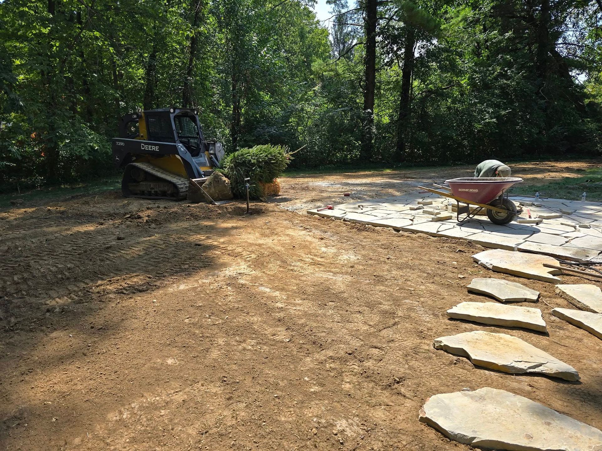 A skid steer, wheelbarrow, and stone path are on a dirt area in front of a wooded area.