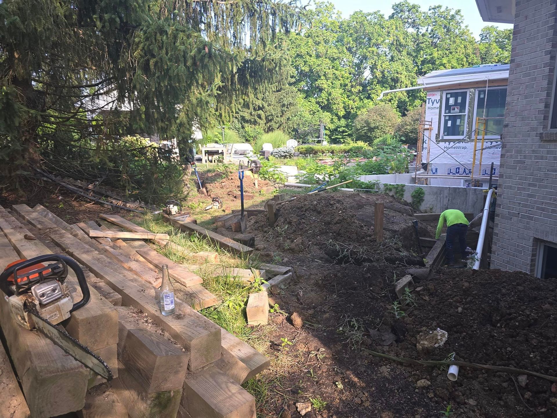 Construction site with a chainsaw, piles of dirt, retaining walls, and a worker in a green shirt.