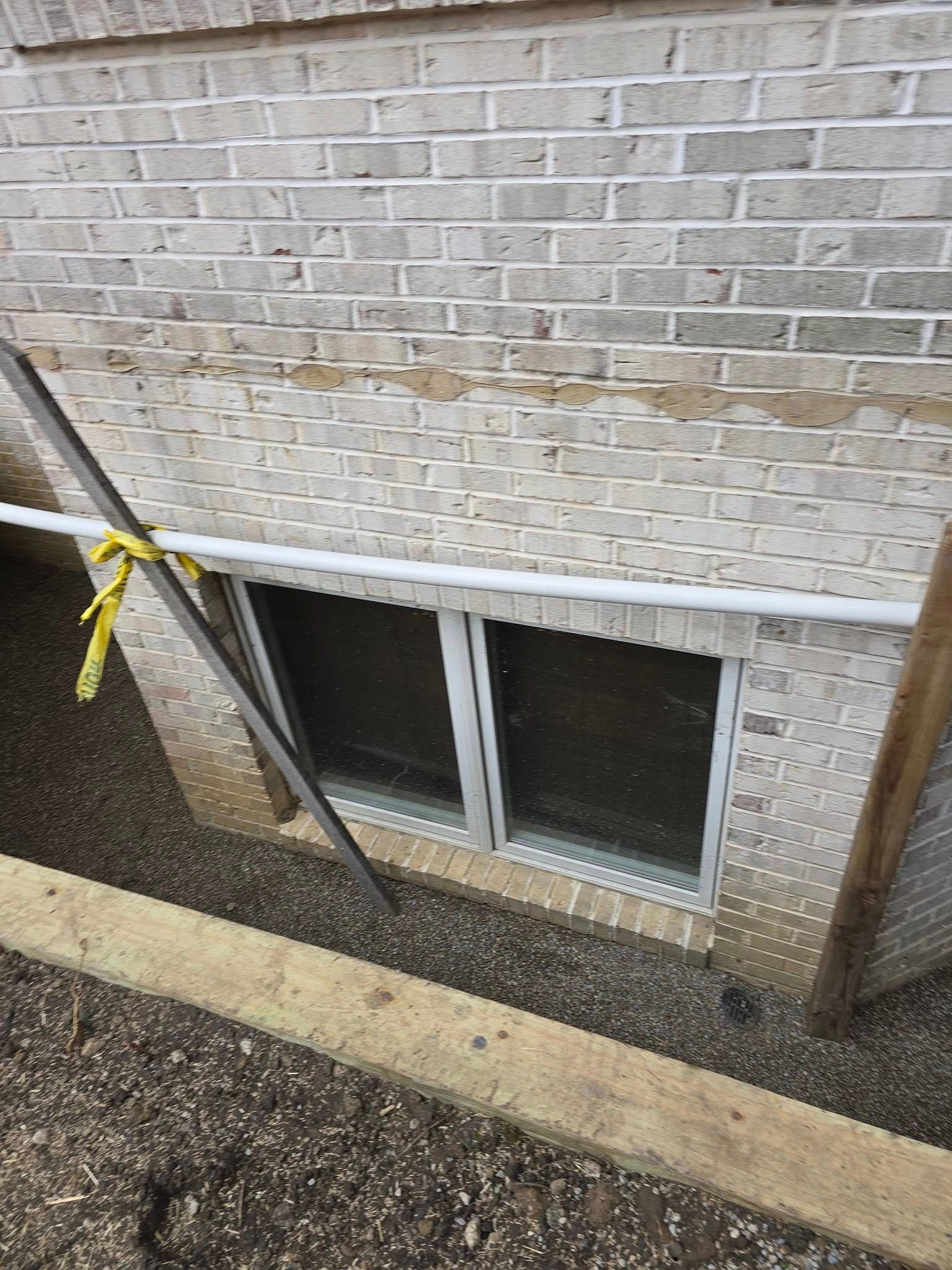 Window well with brick wall above. A white window sits below a white pipe. Yellow caution tape is visible.