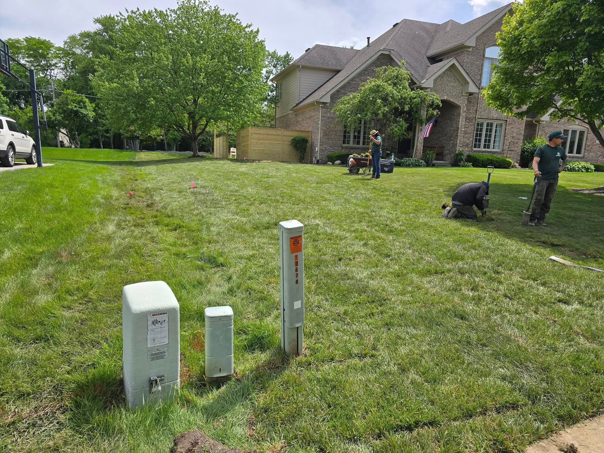 Workers tending a lawn in front of a large house. Green grass, utility boxes, trees, and sky visible.