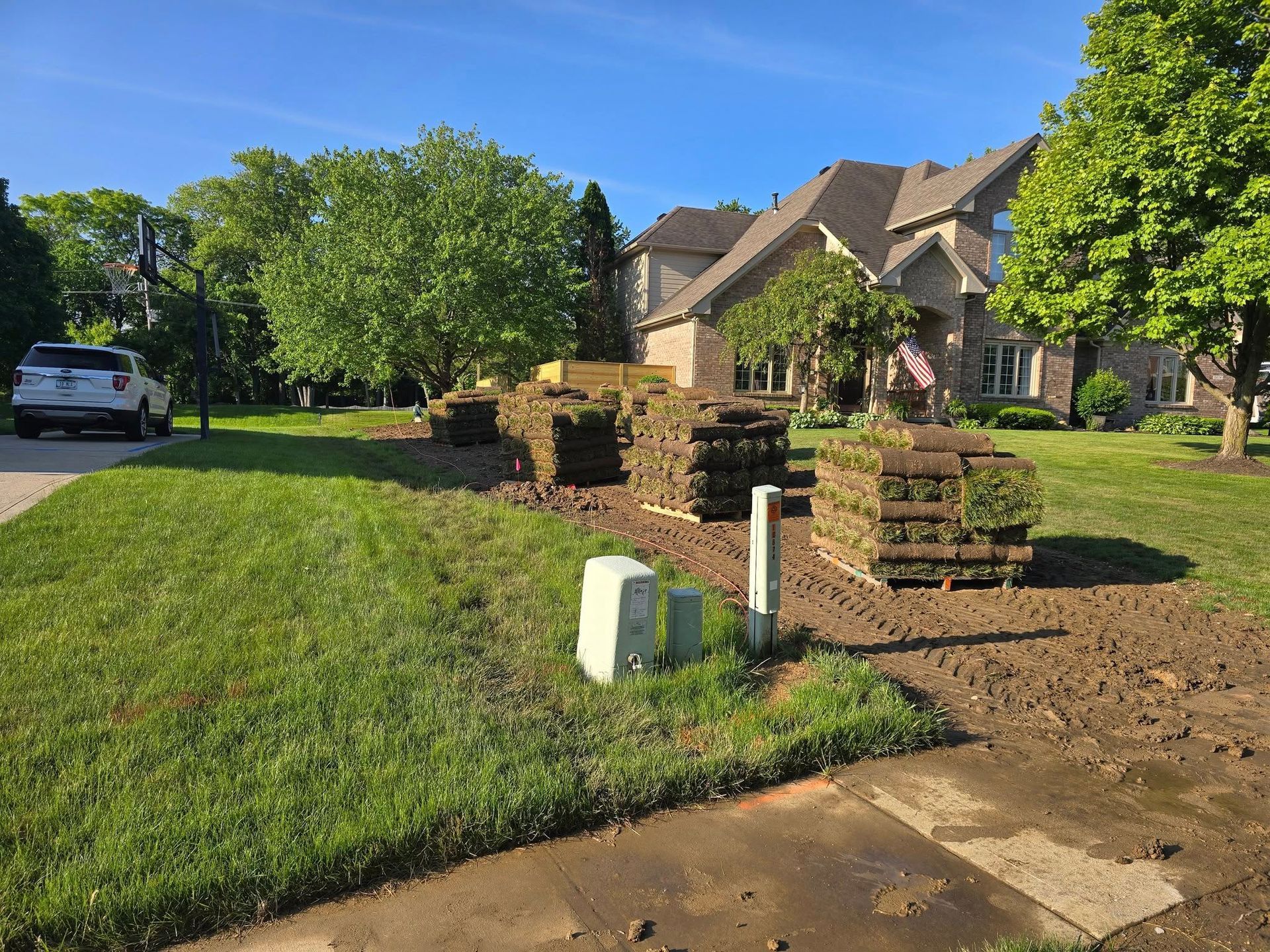 Sod stacked on brown soil in front yard of a two-story home, with a utility box and car present.