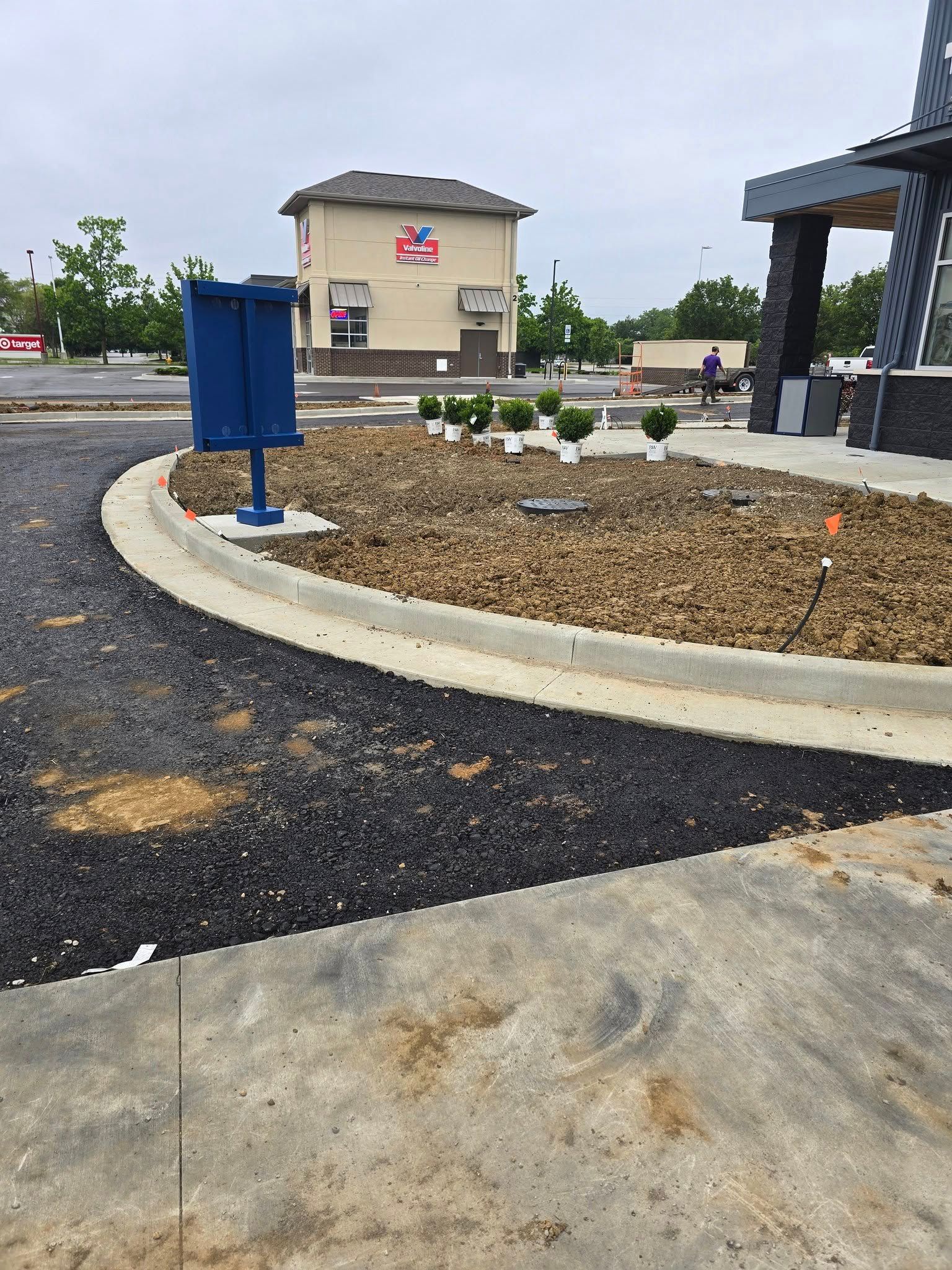 Construction site with curb, dark paving, dirt, and blue mailbox. Building in background.