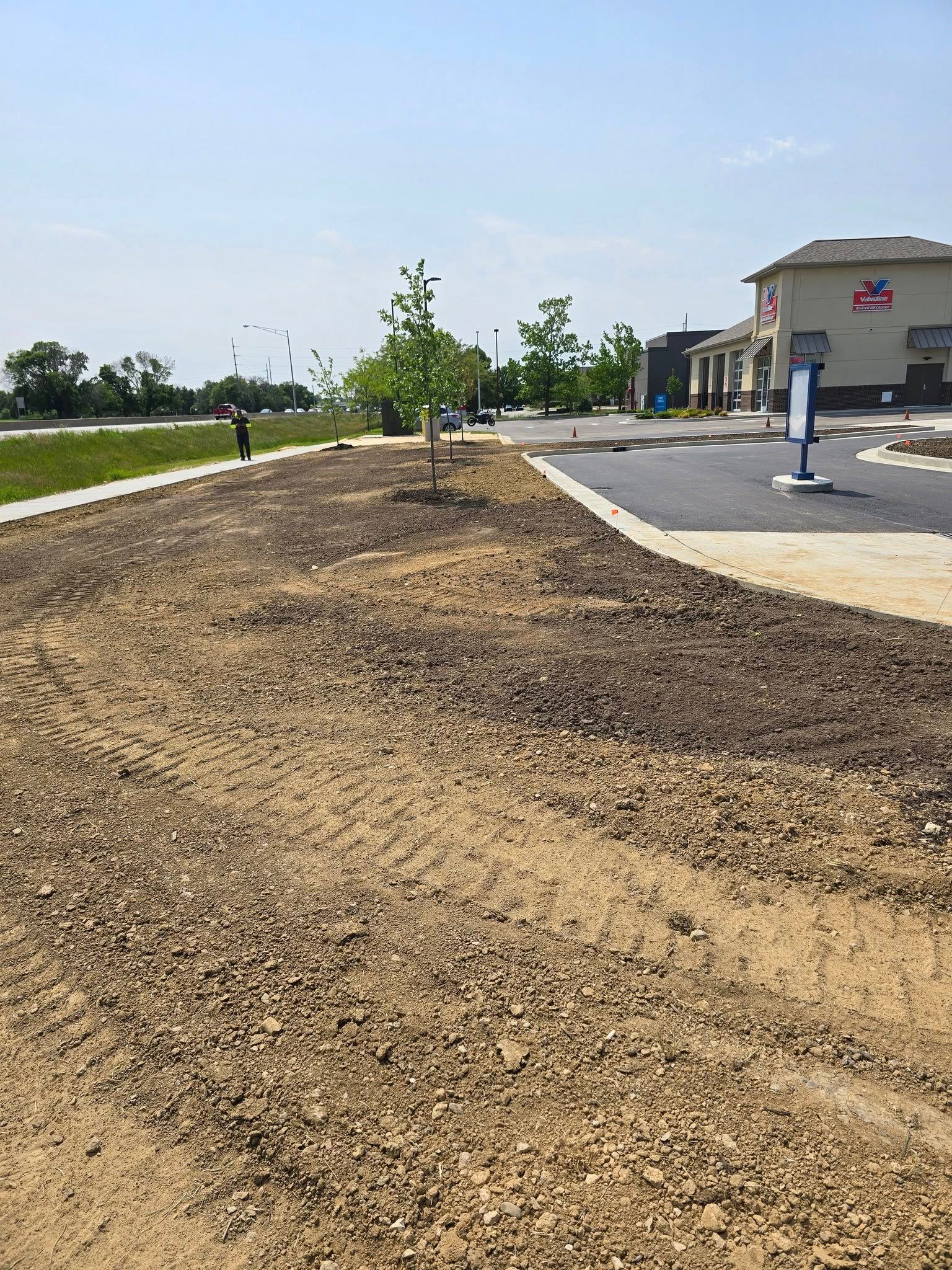 Dirt and gravel landscape with young trees, near a building and paved area. A person stands in the distance.