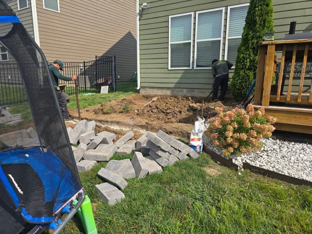 Two workers building a patio. Gray bricks and excavated dirt are in a yard near a house.