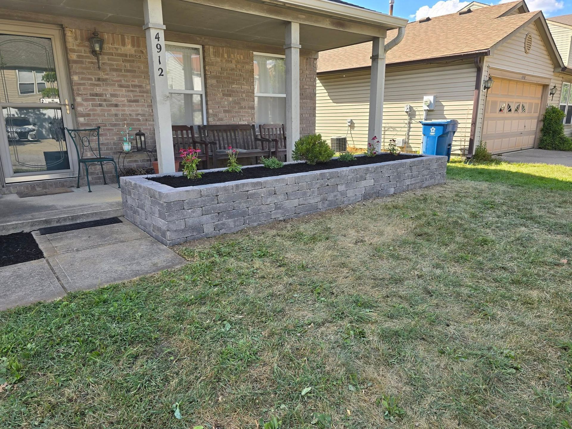 Front of a house with a stone planter box filled with plants, on the lawn. A bench sits on the porch.