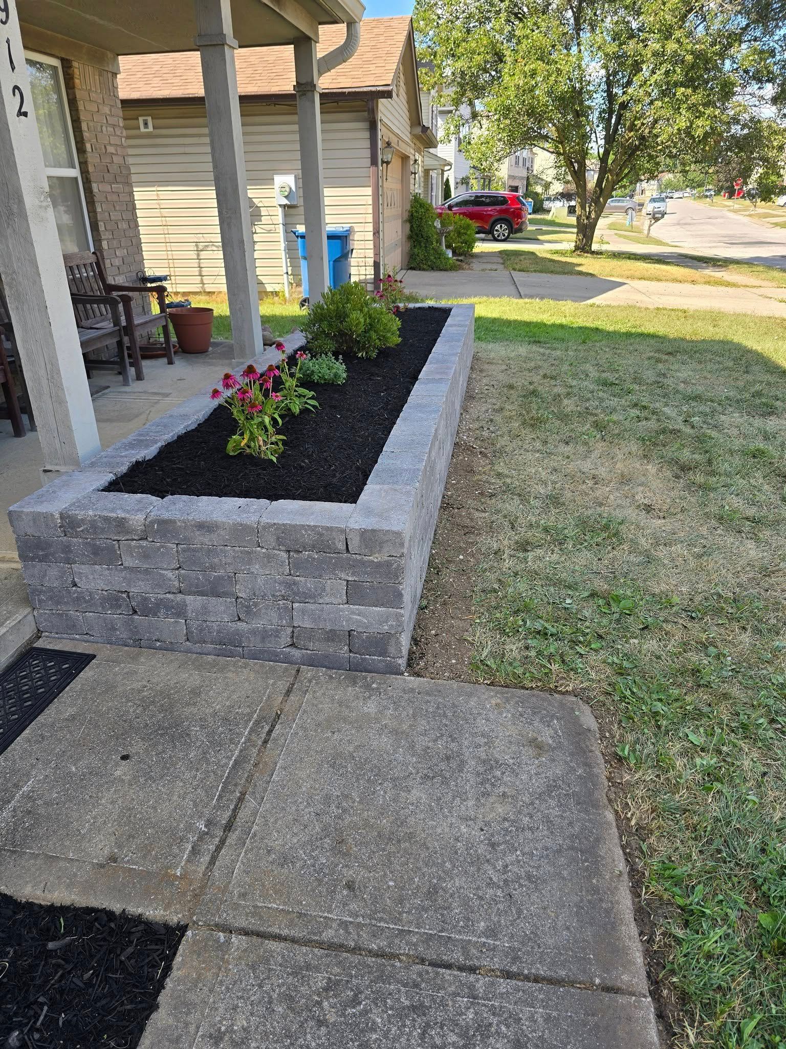 Brick planter with dark mulch and flowers next to concrete walkway and grass lawn.