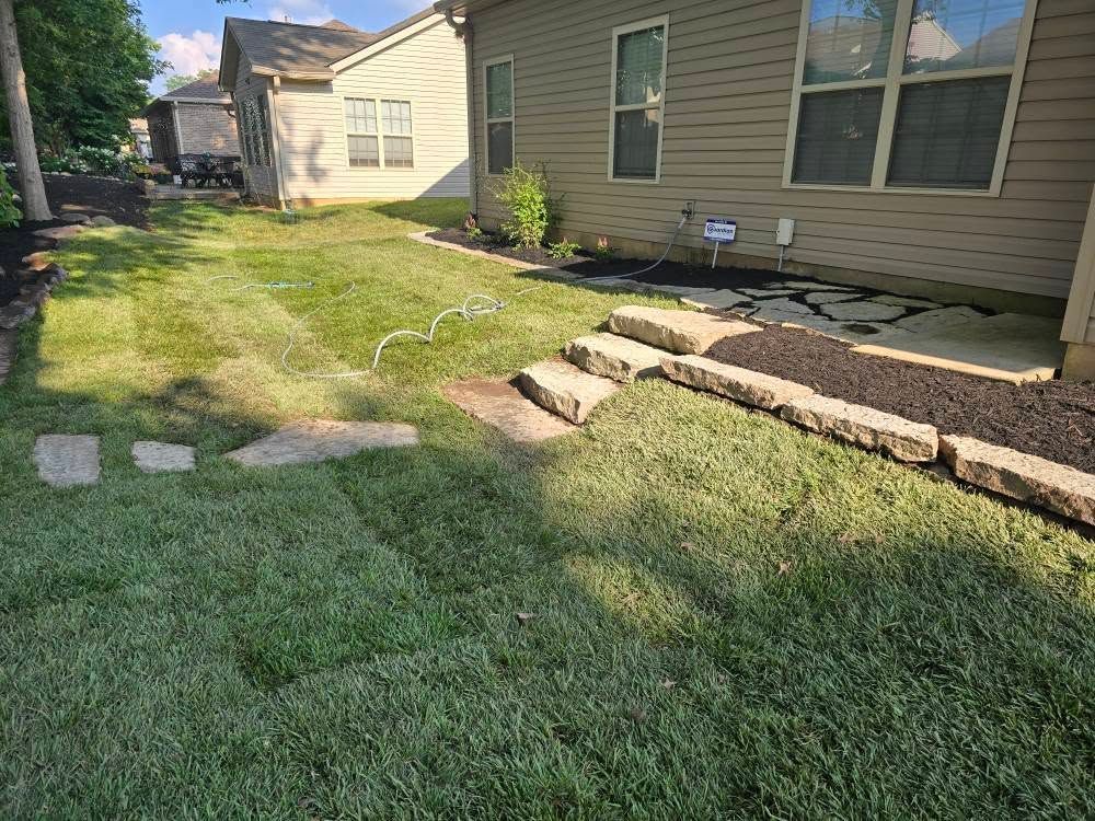 Backyard with green grass, stone steps, and a beige house. Mulched flower beds along the house.