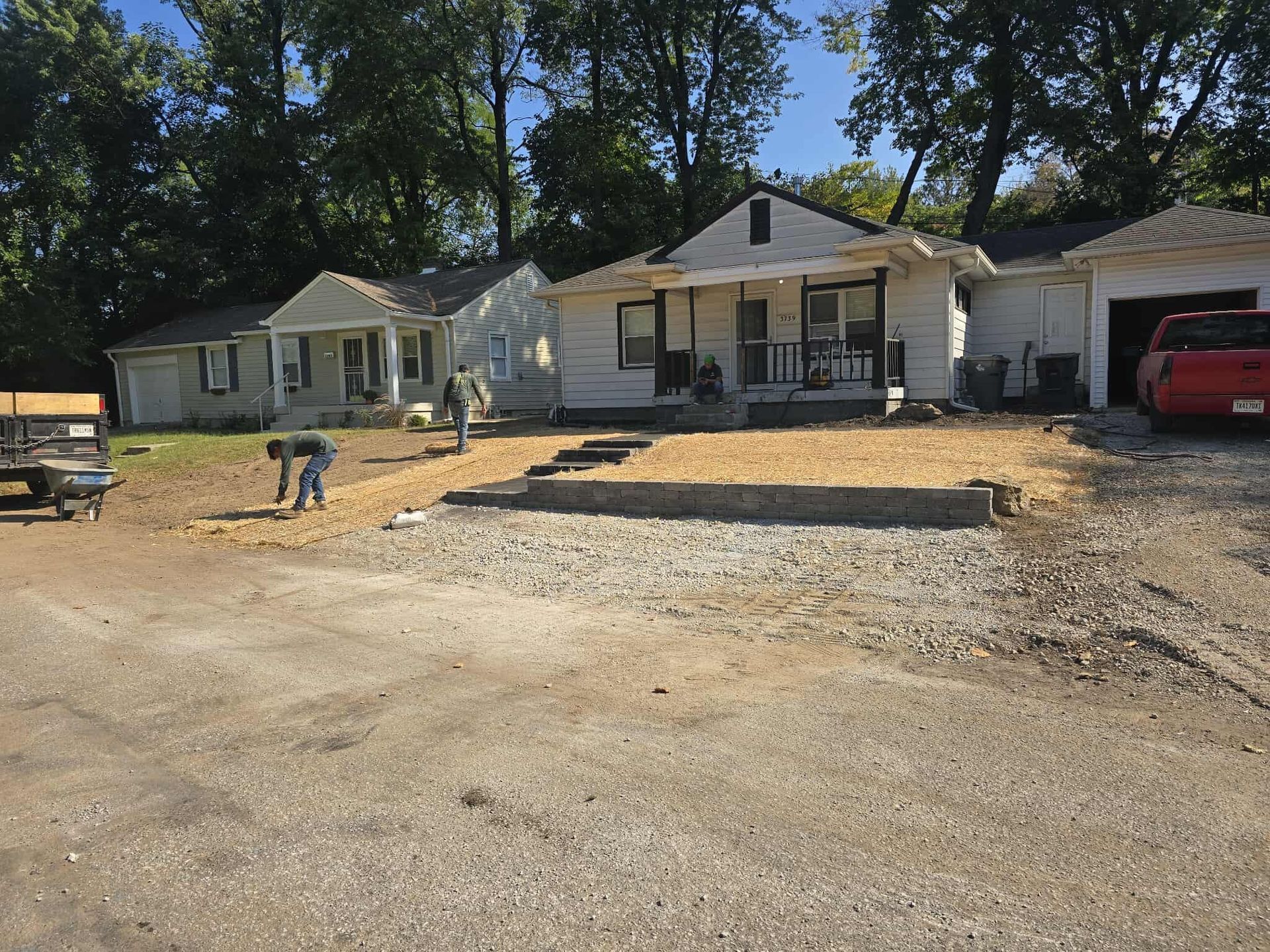 Workers paving a front yard with gravel, in front of a white house with a porch and two other houses.