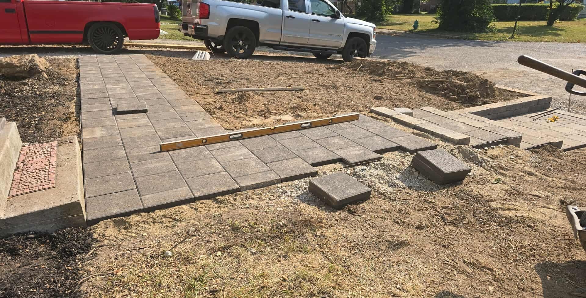 Construction of a brick pathway and patio in a yard, with trucks in the background.
