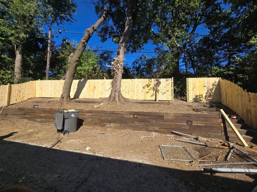 Wooden fence encloses a dirt yard with trees. Gray trash cans and construction debris are visible. Blue sky in the background.