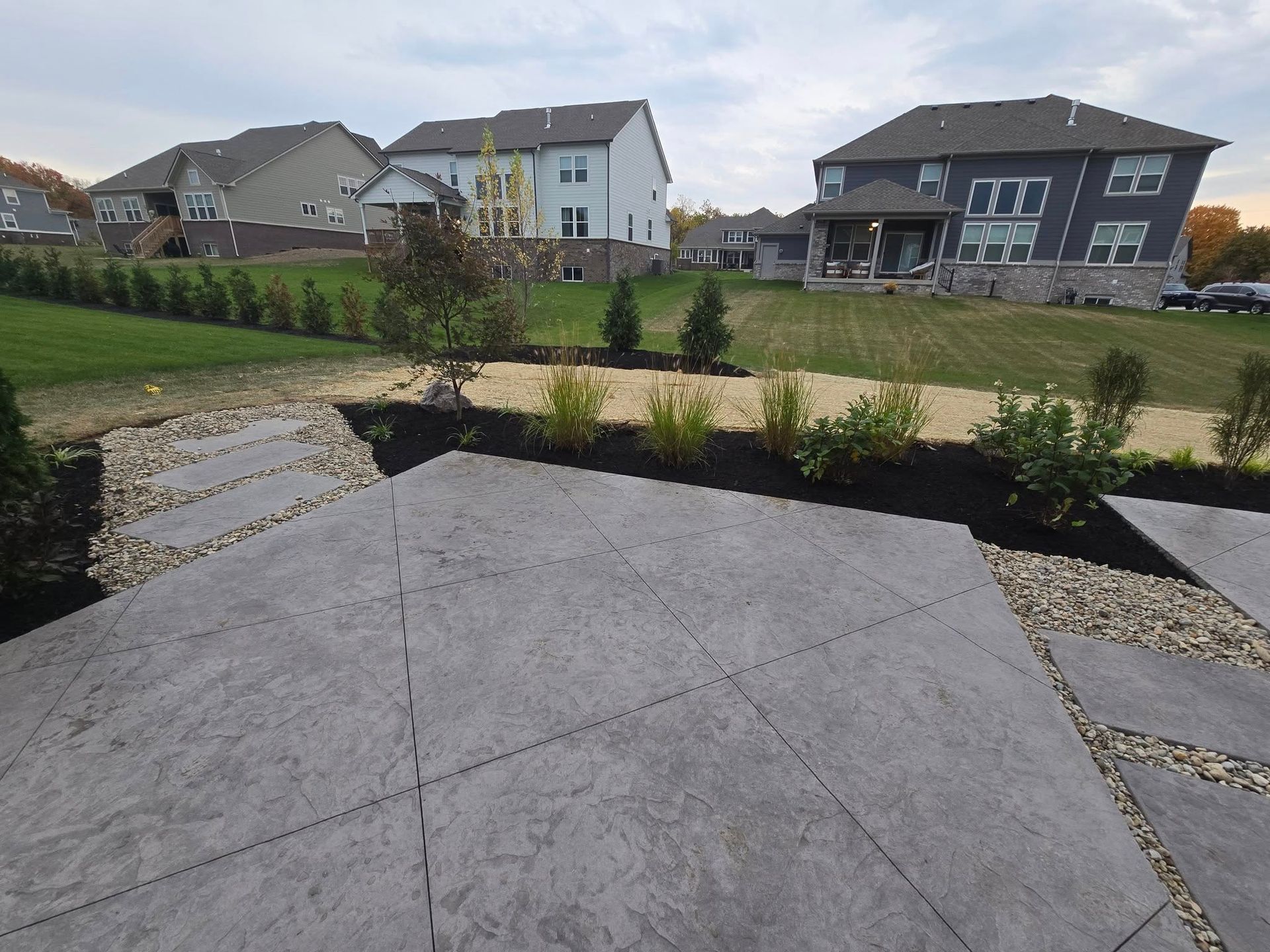 Concrete patio with landscaping, small trees, and houses in the background on a cloudy day.