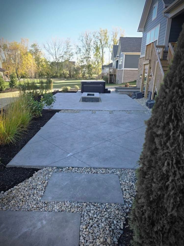 Concrete patio with a fire pit, bordered by rock and landscaping, in front of houses.