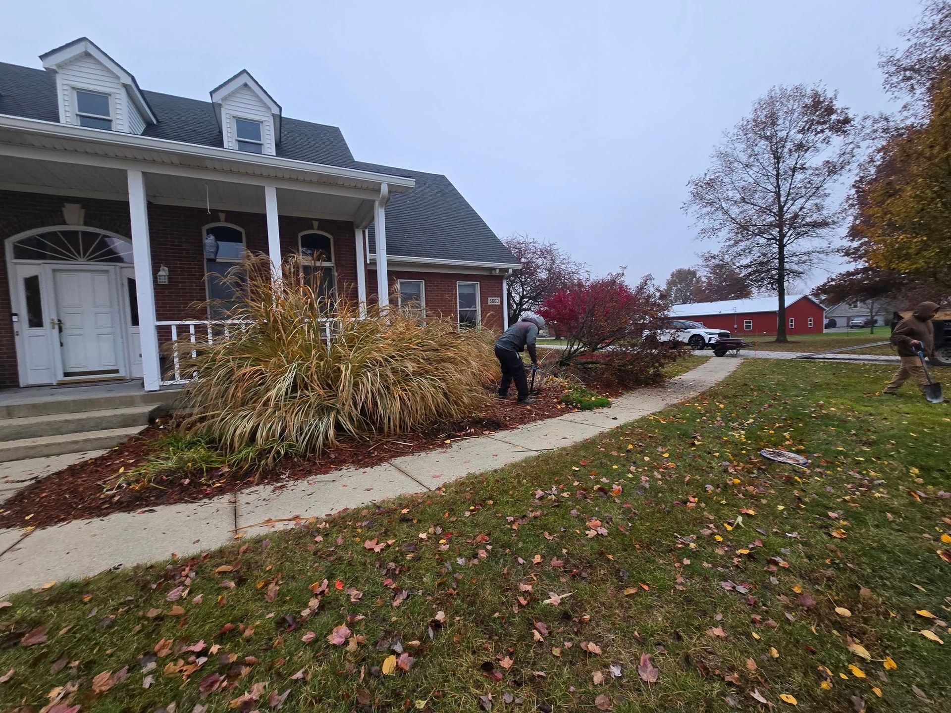 Person tending to plants in front of a brick house with white trim; autumn leaves on the ground.