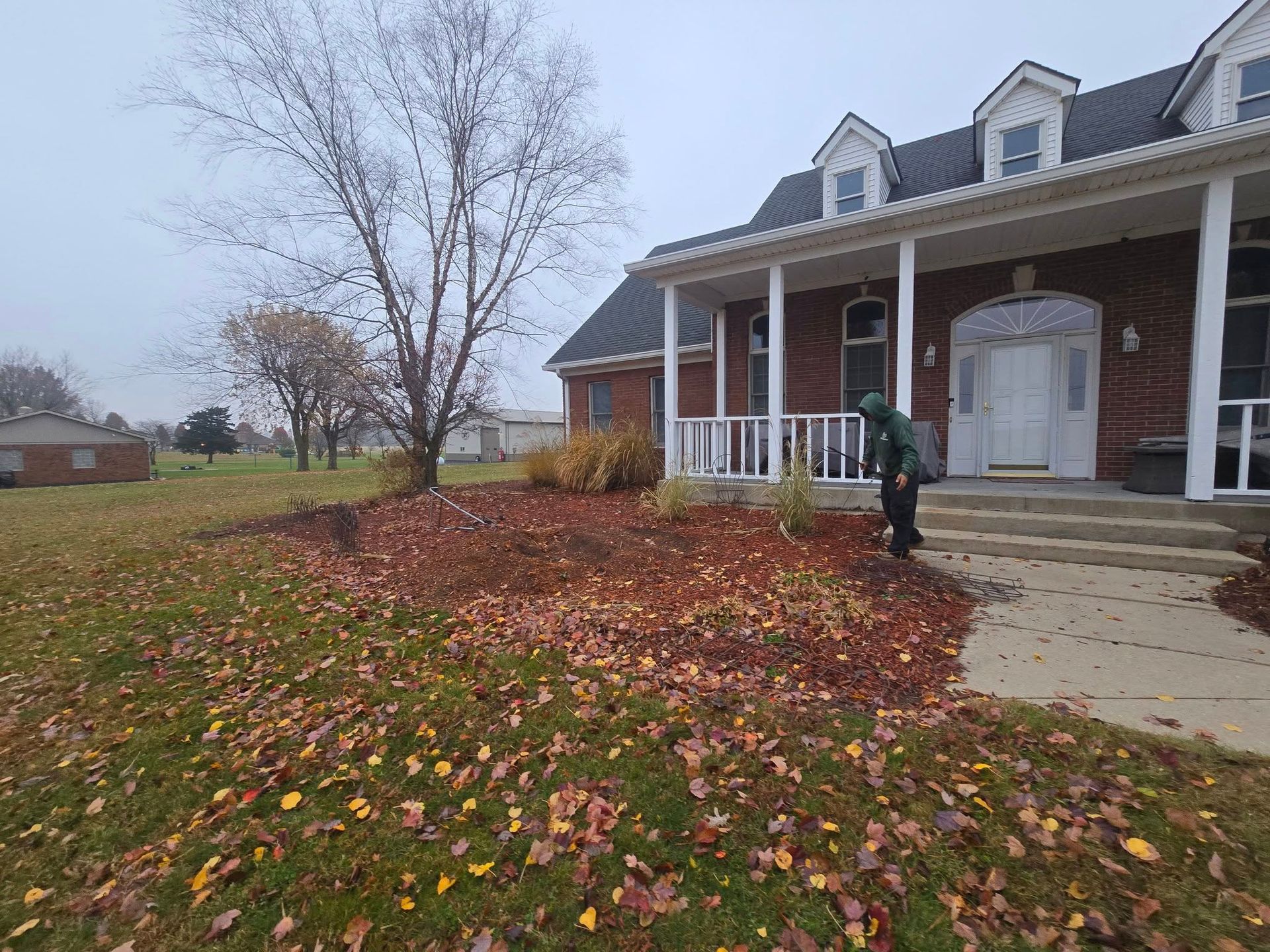 Person raking autumn leaves on the lawn in front of a brick house with a white porch on an overcast day.
