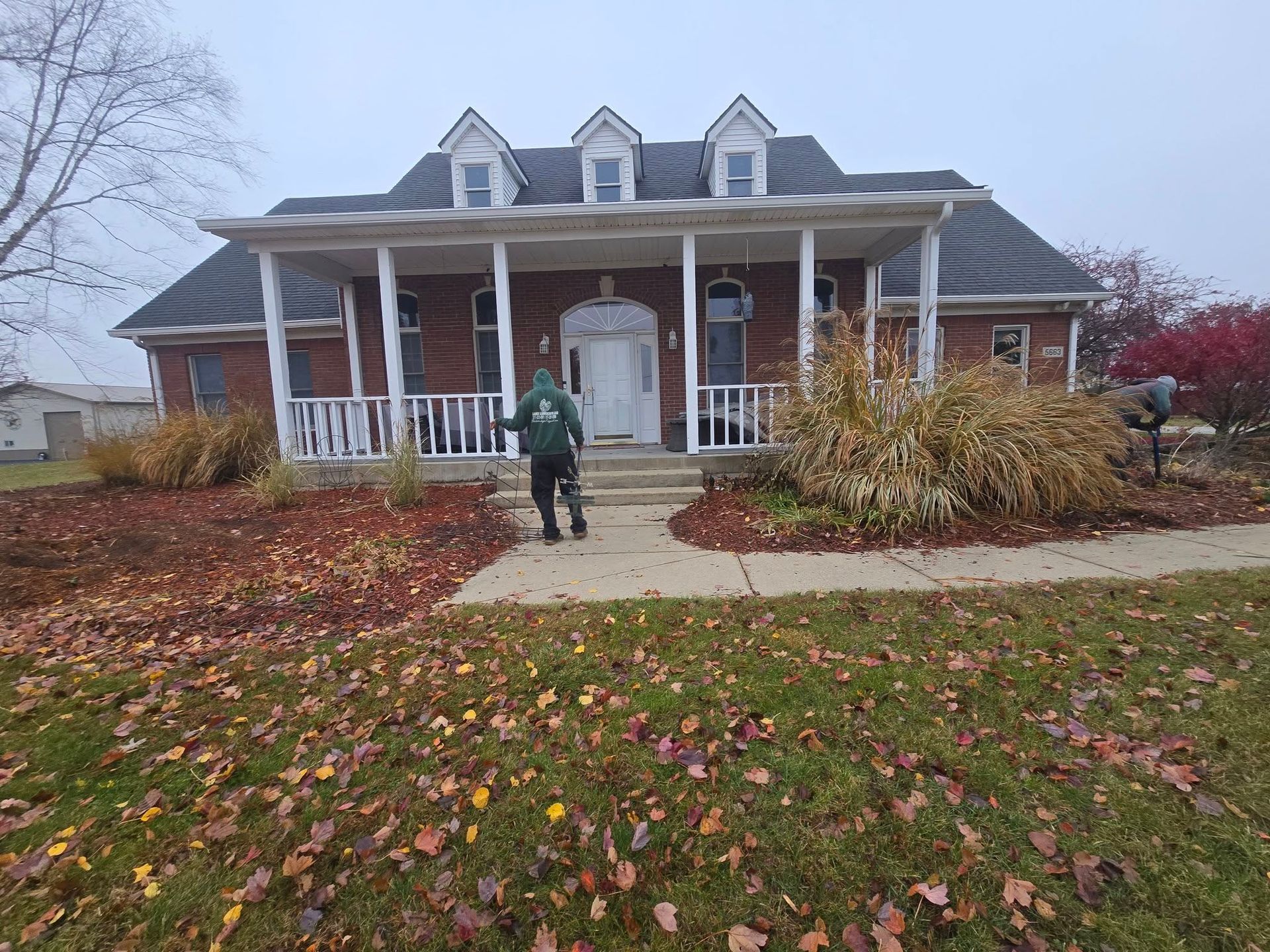 Person standing in front of a brick house with white porch and fall foliage.