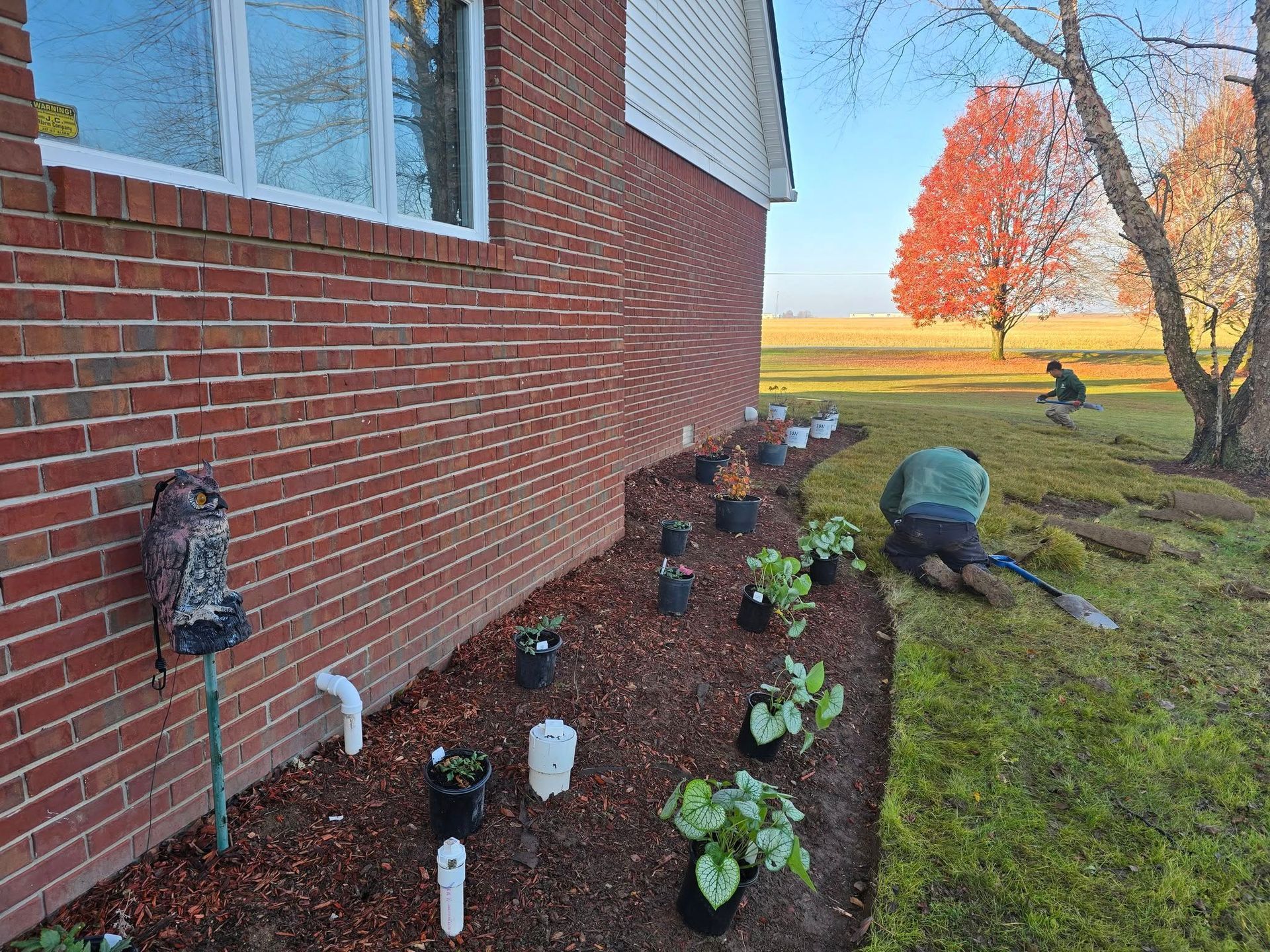 Two people planting flowers along a brick house. Fall foliage and field in the background.