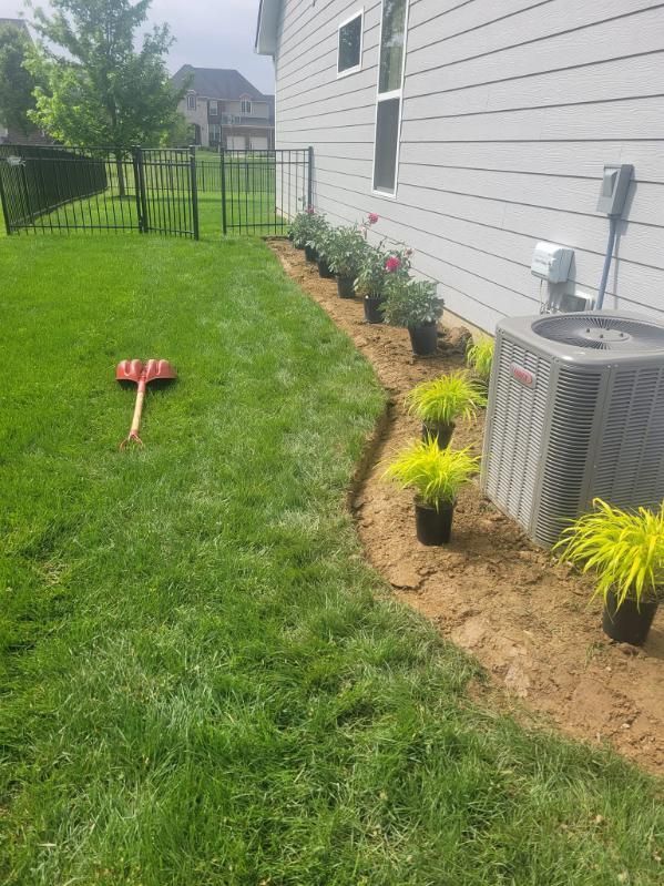 A residential backyard with newly planted flowers and grass, next to a gray house, with an AC unit and a rake.