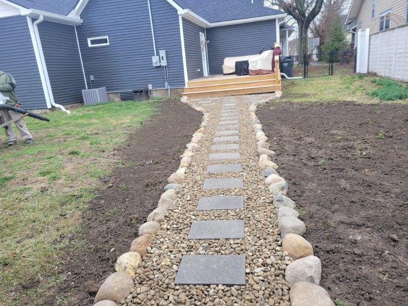 Stone path through yard leading to a wooden deck, bordered by rocks and gravel, next to a house.
