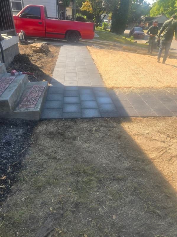 A walkway under construction; gray pavers, gravel, red truck, workers, and grass.