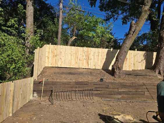 Wooden fence built around a yard on a sloped terrain with retaining walls and trees.