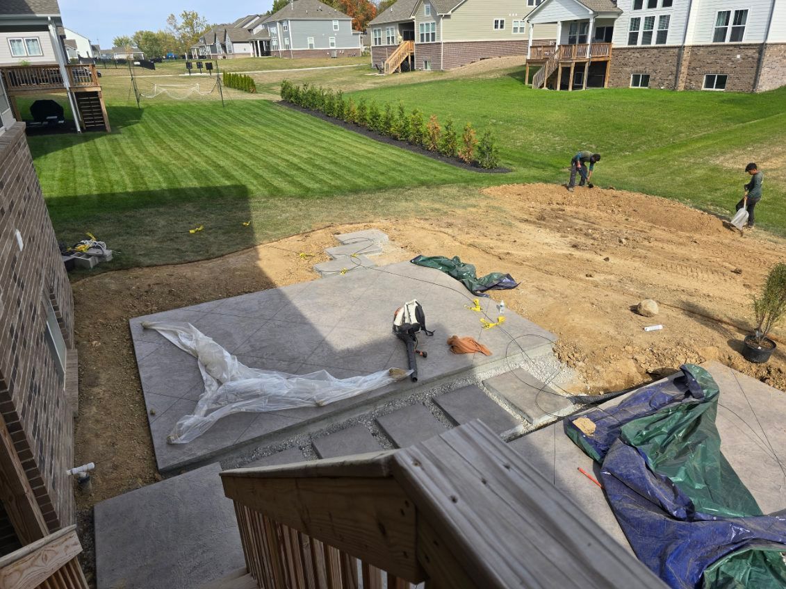 Construction site with concrete patio, steps, and workers in a residential backyard. Green lawn, trees, and houses in the background.
