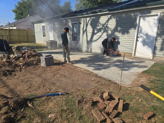 Two people building a patio. One cuts stone, the other stands. Dirt, stone, and grass surround the project near a building.