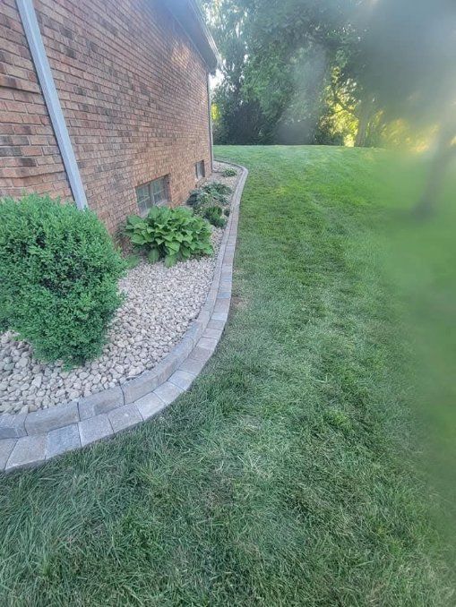 A brick building with a landscaped bed of plants and rocks, bordered by paving stones, next to a grassy lawn.