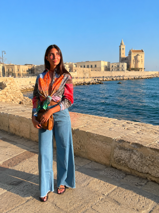 Una donna in piedi accanto a un muretto di pietra sul lungomare, con indosso una blusa colorata e jeans, sullo sfondo un lungomare storico.