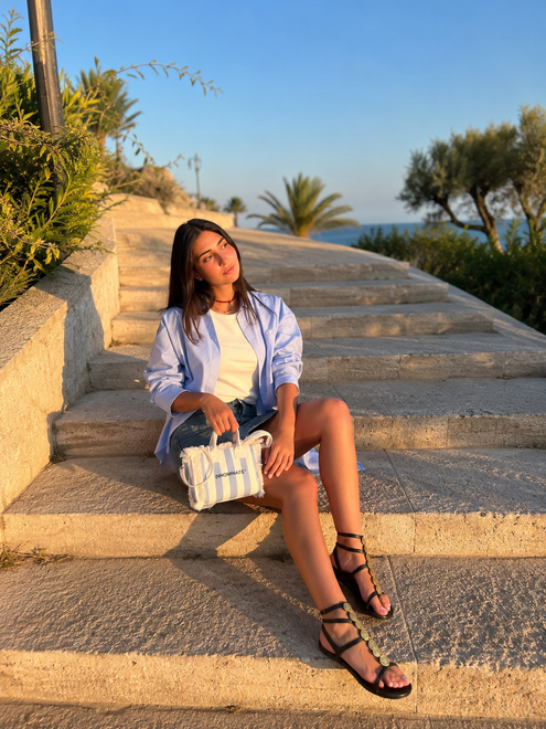Donna seduta su gradini di pietra illuminati dal sole in riva al mare, indossa una camicetta chiara e sandali neri con una borsa di paglia.