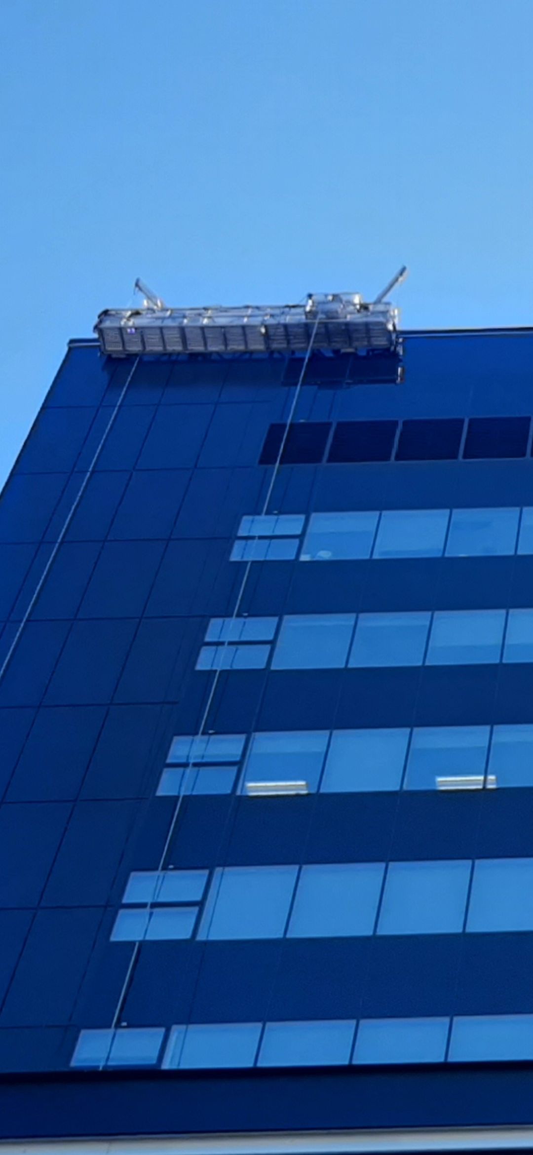 Une plateforme de maintenance suspendue à un grand immeuble en verre bleu.