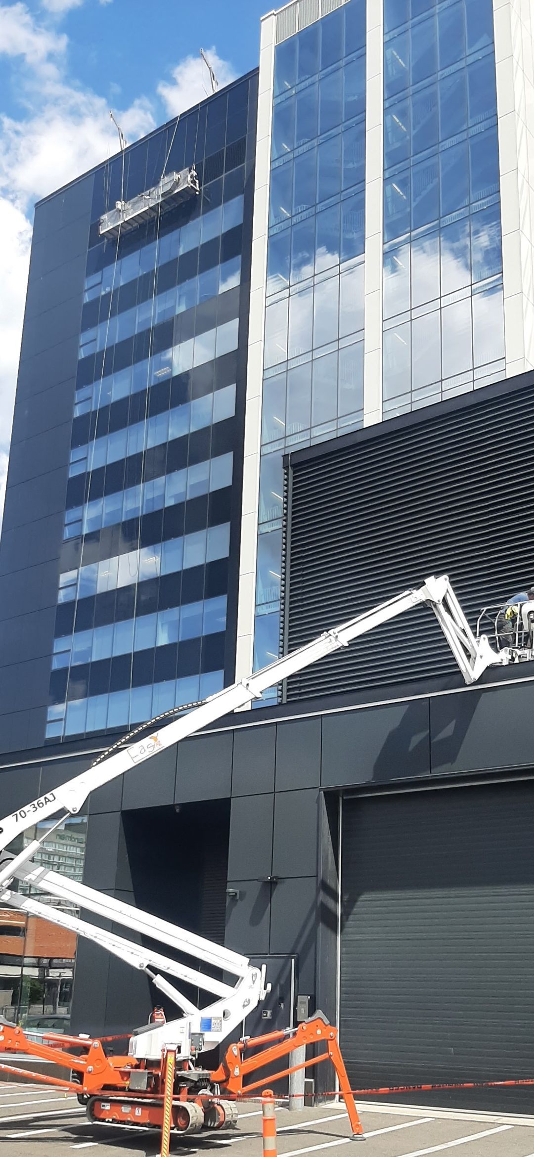 Un immeuble de grande hauteur en verre avec une nacelle élévatrice en position de travail. La nacelle est à l'œuvre sur la façade sombre d'un bâtiment plus bas. Ciel bleu.