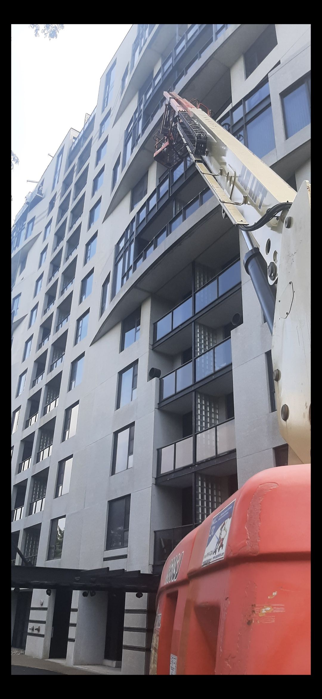 Un monte-charge orange sur un chantier, près d'un immeuble moderne de grande hauteur, doté de nombreuses fenêtres et de balcons.