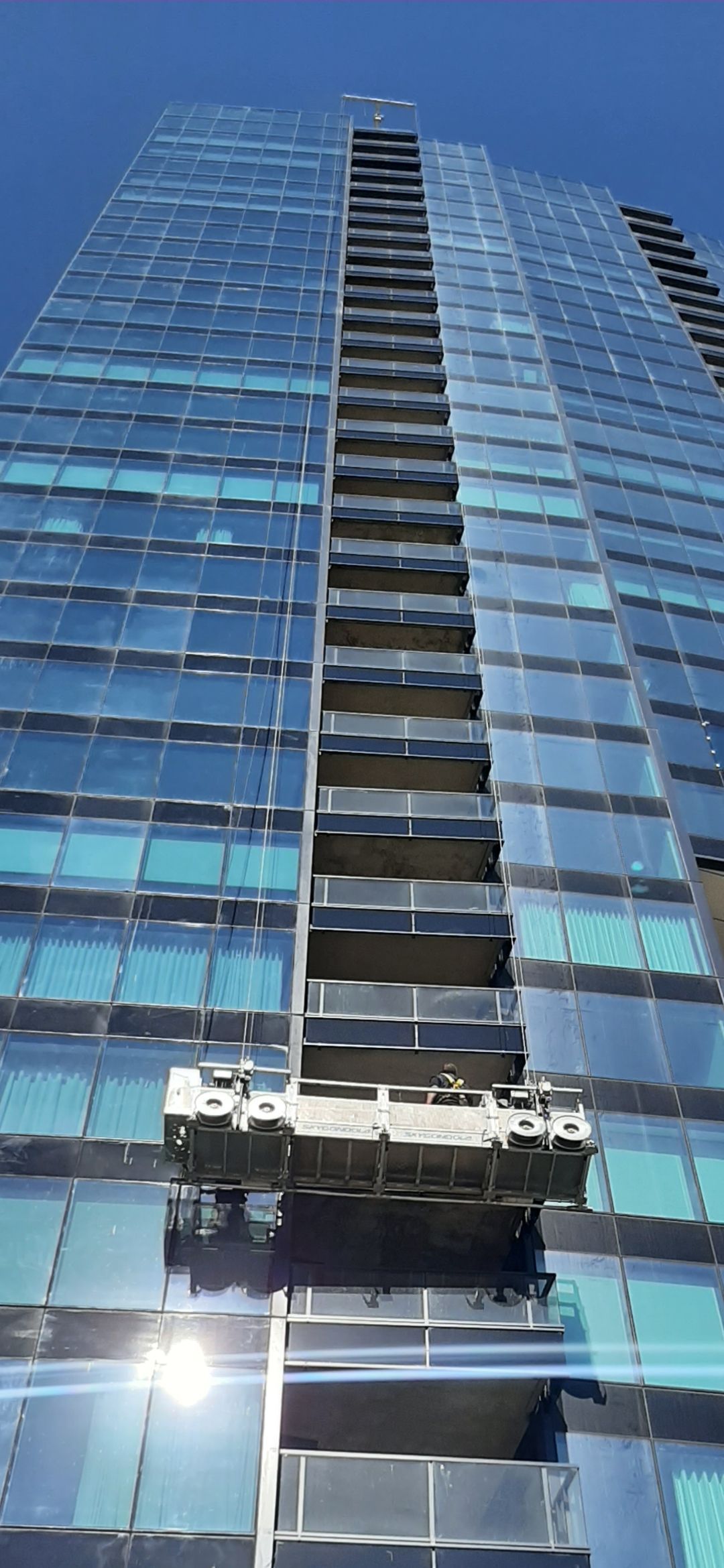 Vue en contre-plongée d'un gratte-ciel en verre doté d'une plateforme de lavage de vitres. Ciel d'un bleu éclatant.