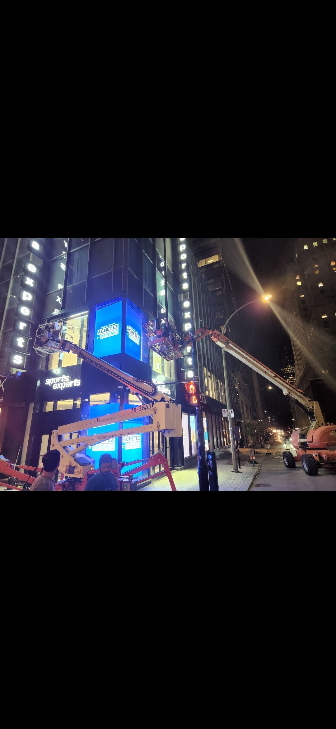 Des ouvriers du bâtiment se trouvent sur des plateformes élévatrices près d'un bâtiment aux enseignes lumineuses, de nuit.