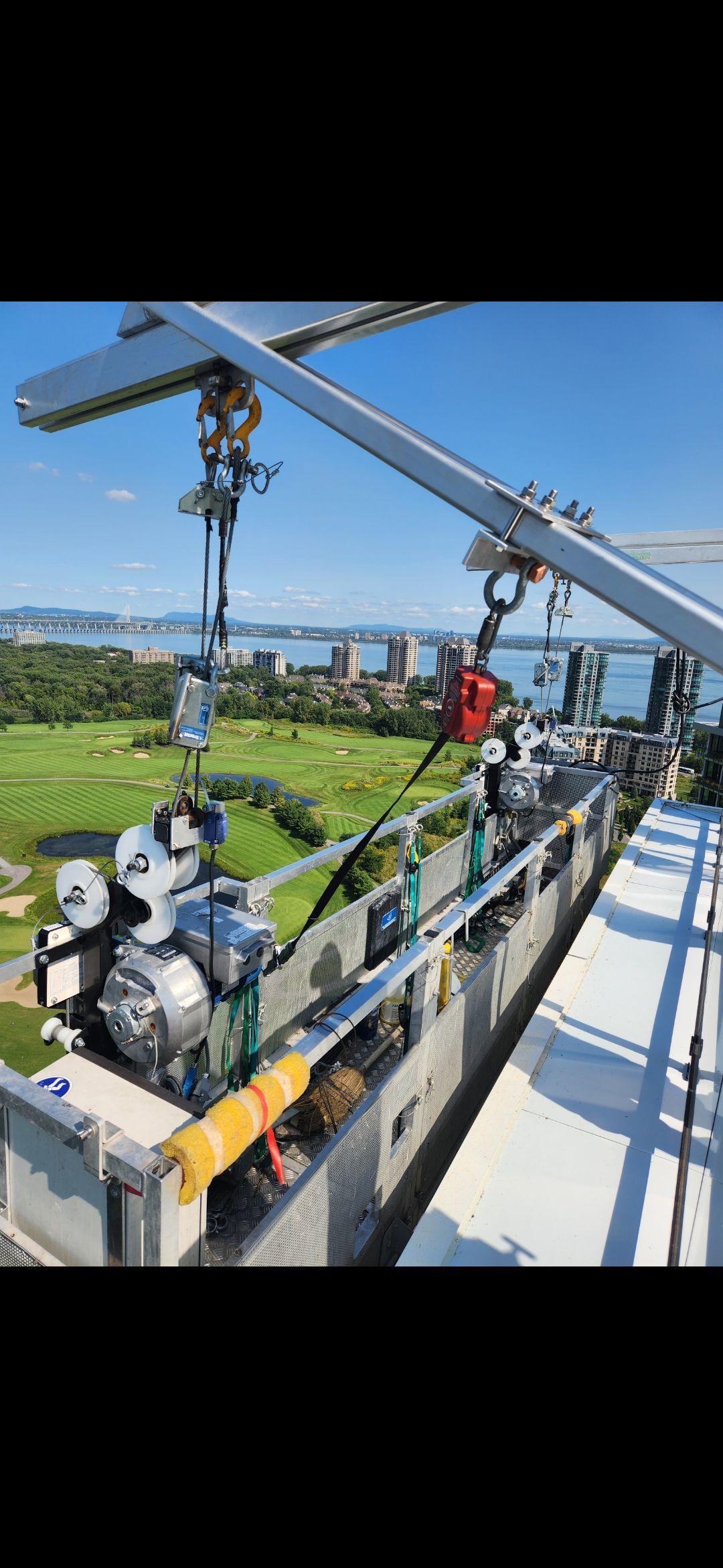 Une grue de chantier sur un immeuble de grande hauteur, avec ses engins et une vue sur la verdure et l'eau.