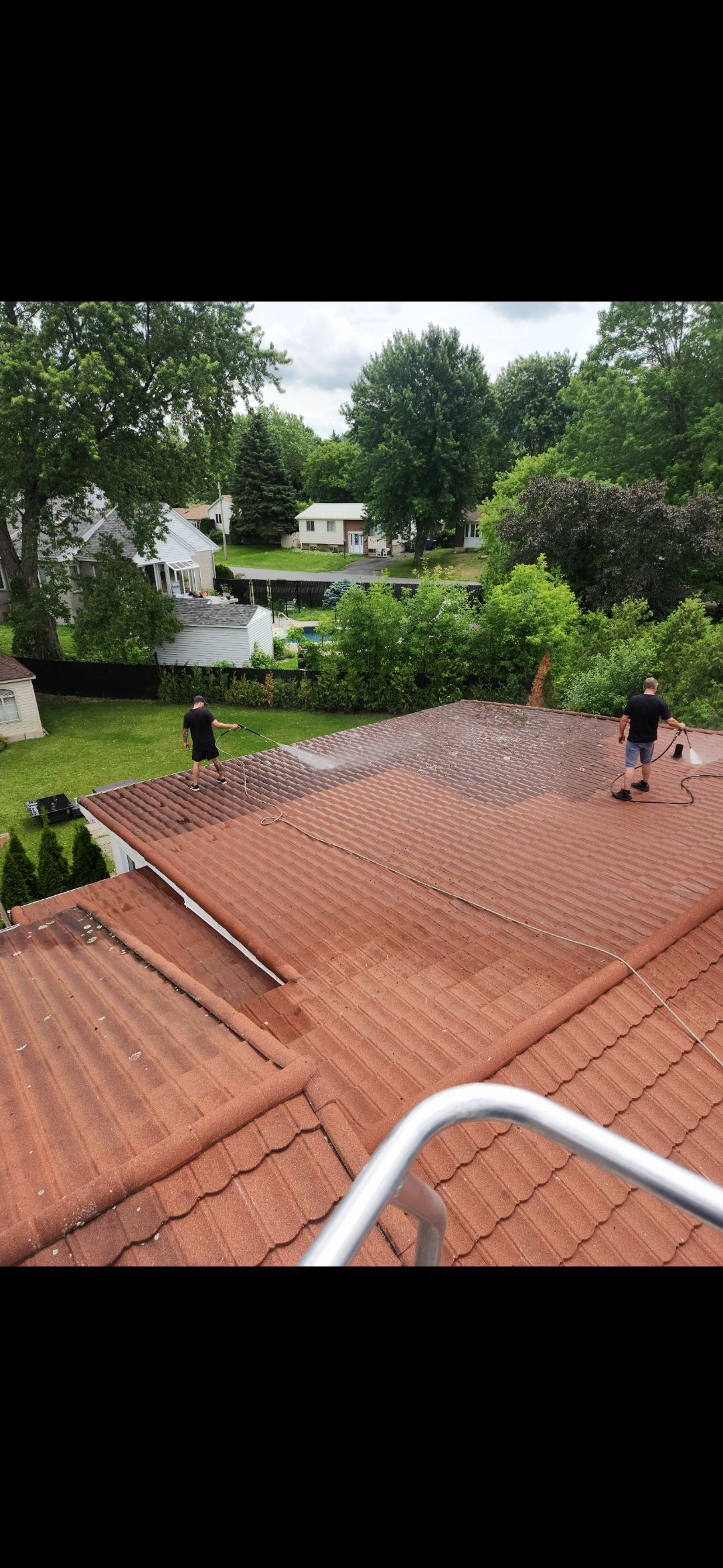 Deux personnes travaillent sur un toit de tuiles en terre cuite, entourées d'arbres verdoyants et de maisons, par une journée nuageuse.