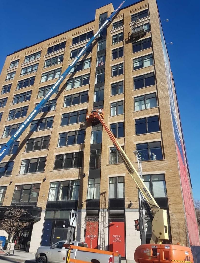 Des ouvriers du bâtiment travaillent sur des plateformes élévatrices, sur la façade d'un grand immeuble en briques, par une journée ensoleillée.