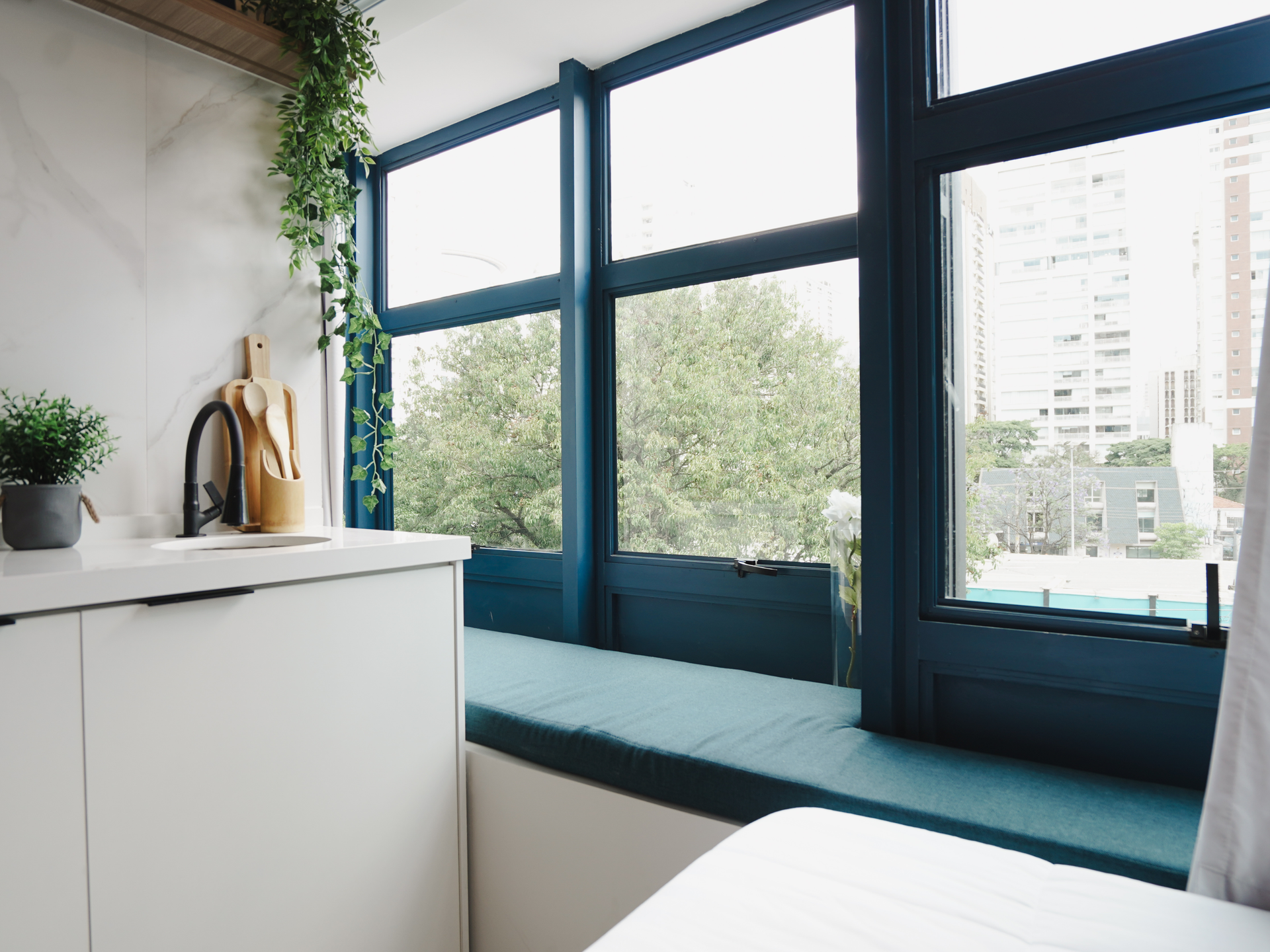 Quarto moderno e luminoso com janelas amplas, almofada de banco azul-petróleo, armário branco e plantas em vasos.