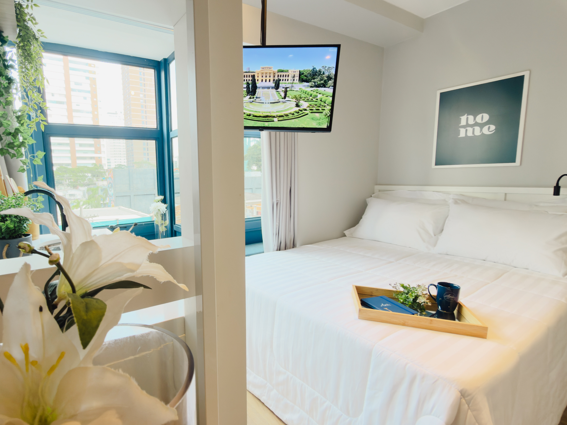 Quarto branco e luminoso com janelas amplas, cama, TV fixada na parede e bandeja com flores sobre a cama.