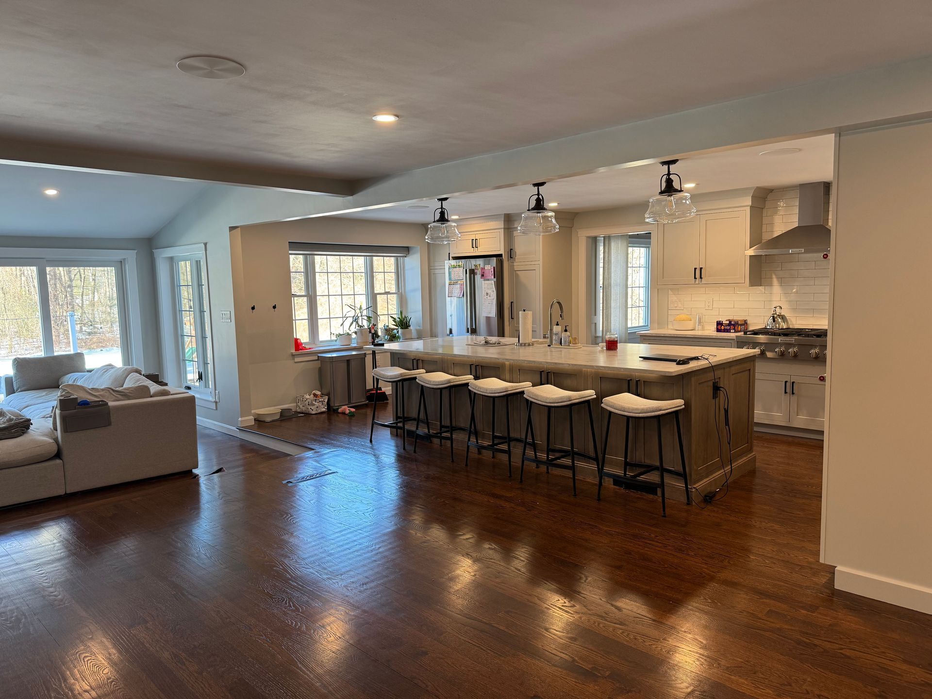 An open-concept room features a kitchen with an island, bar stools, and hardwood floors, leading into a living area.