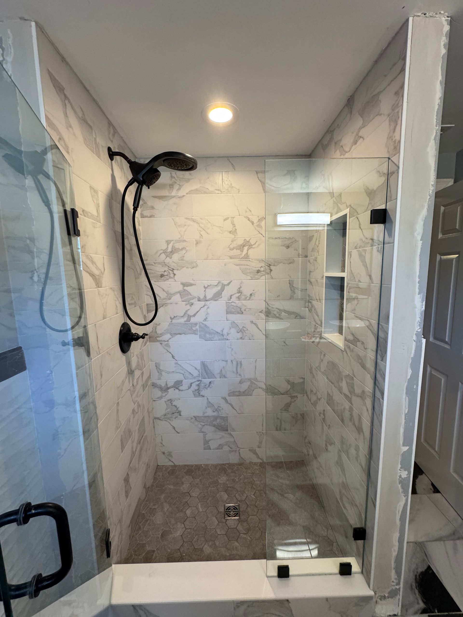 A modern walk-in shower with white marbled tile walls, a dark stone floor, glass doors, and a black rainfall showerhead.