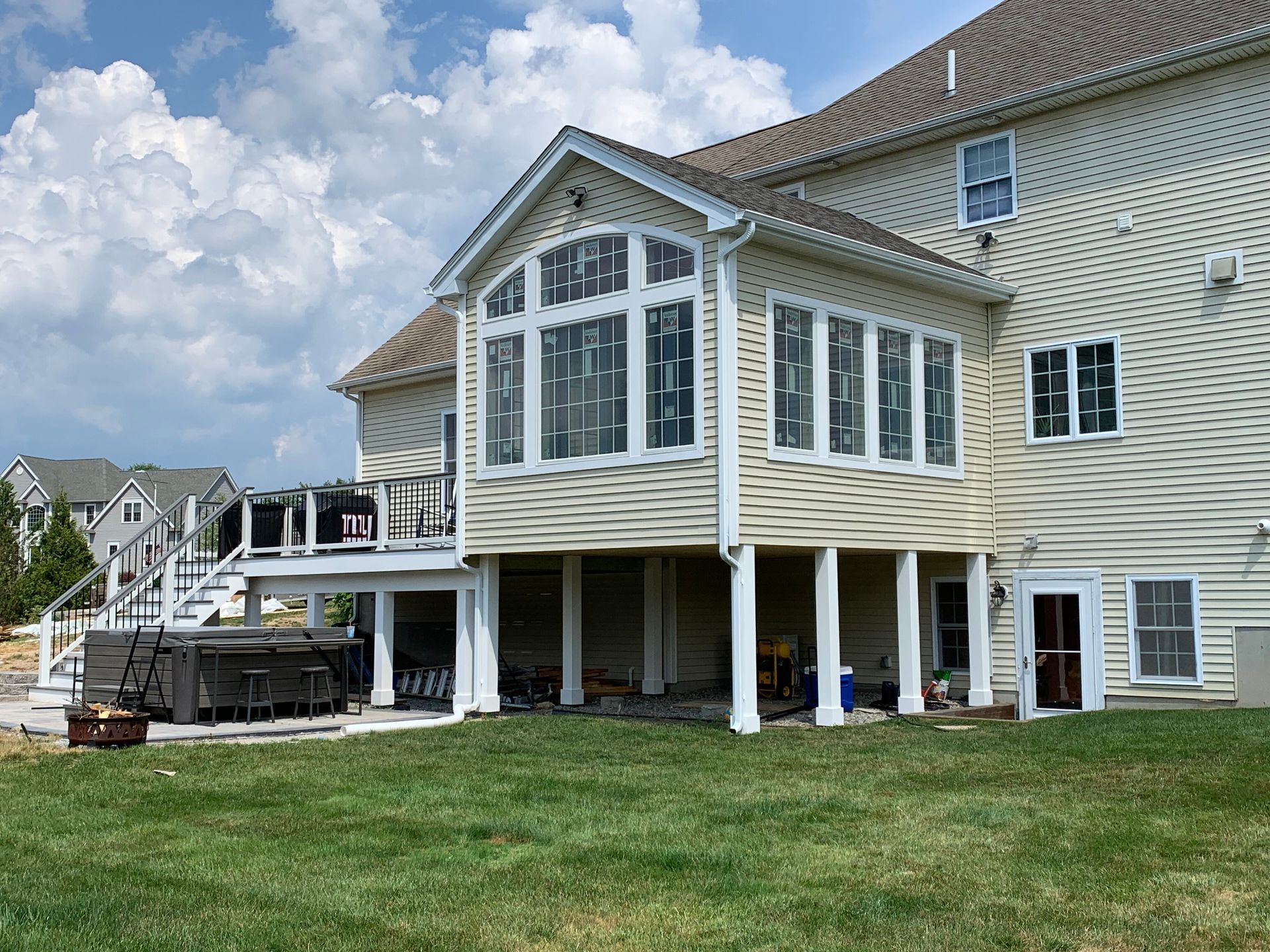 A beige house with a sunroom addition, a raised deck with white railings, and a hot tub on the patio below.
