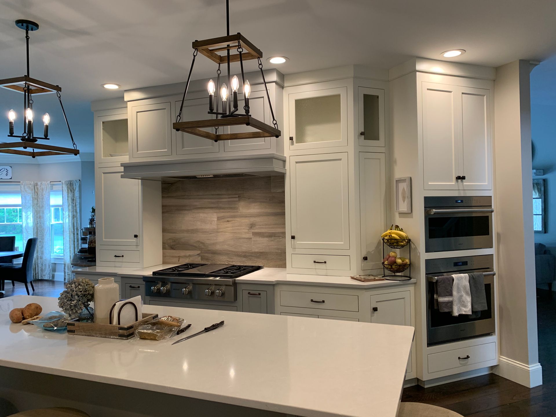 A white kitchen featuring a large island, stainless steel wall ovens, a wood-tiled backsplash, and lantern-style lighting.